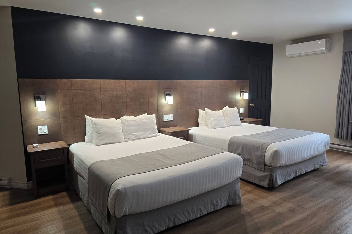 A hotel room with two beds, nightstands, wall-mounted lights, an air conditioning unit, and a wooden floor.