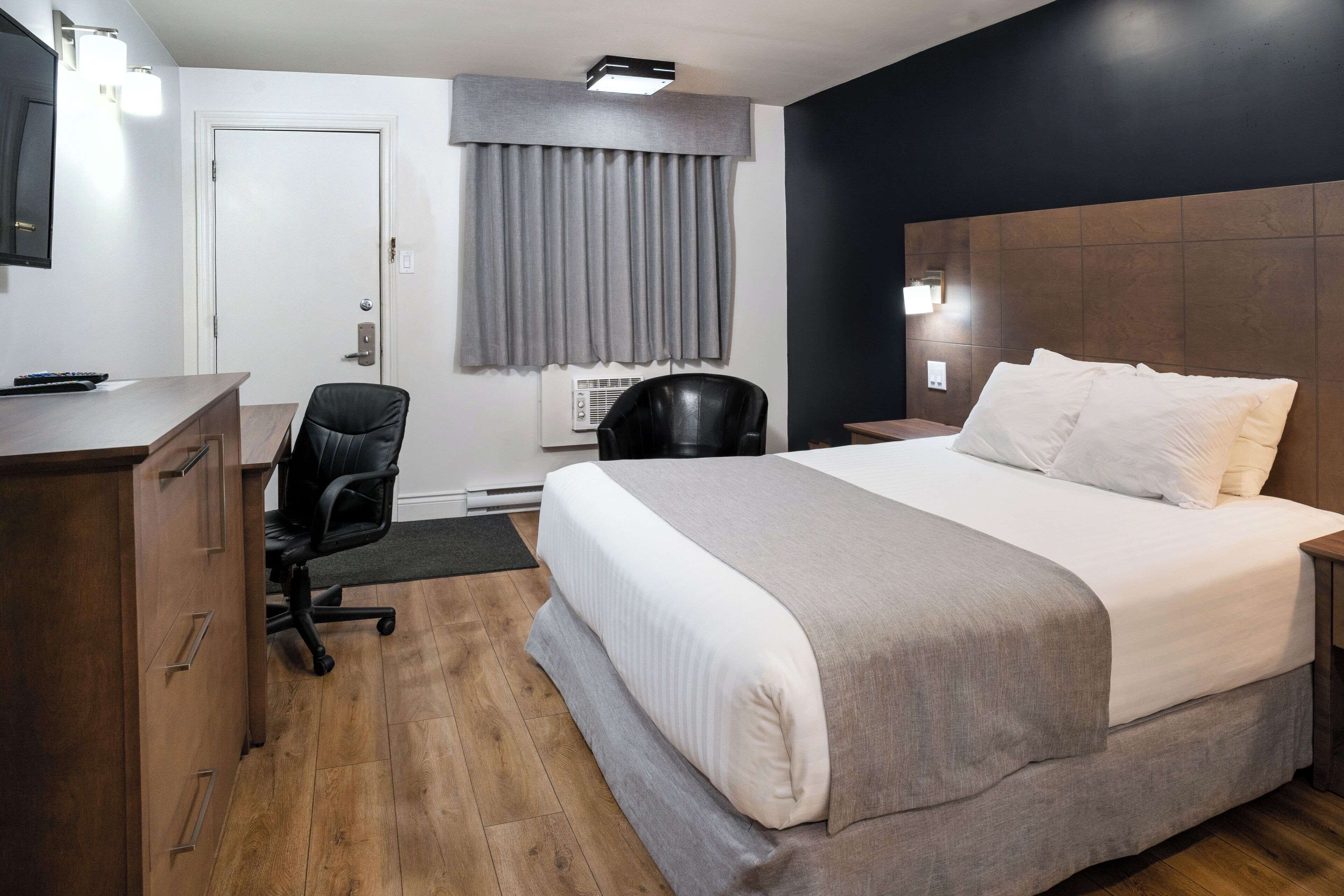 Ein Hotelzimmer mit einem großen Bett, einem Schreibtisch mit Schubladen, einem Sessel, einem Fernseher und einem Fenster mit Vorhängen.