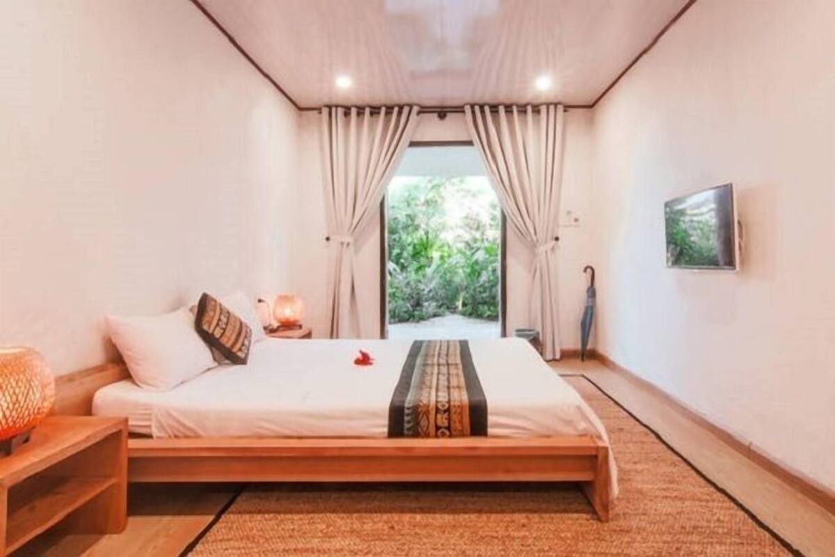 A bedroom with a large bed, a bedside table, a nightstand, a lamp, and a view of greenery outside.