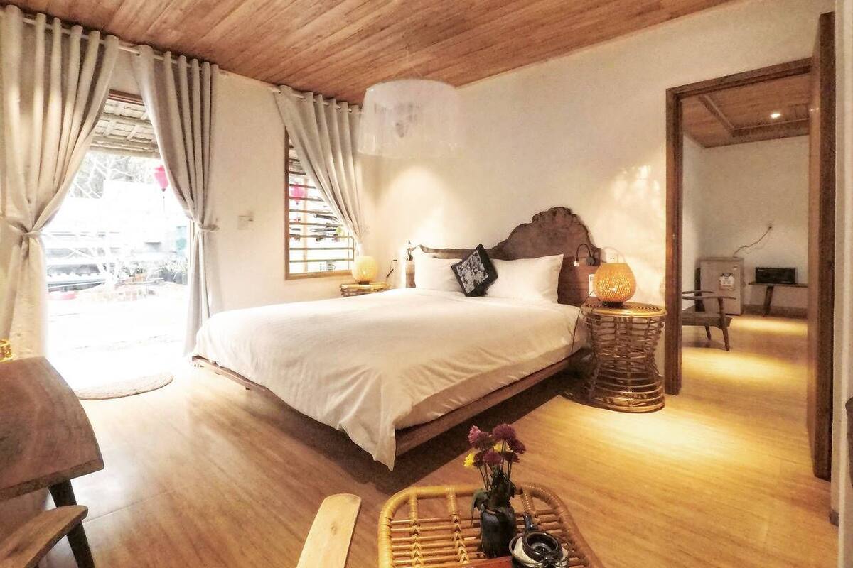 A bedroom with a wooden ceiling, a bed with white bedding, a bedside lamp, a wicker side table, and a view of the outside through a window with curtains.