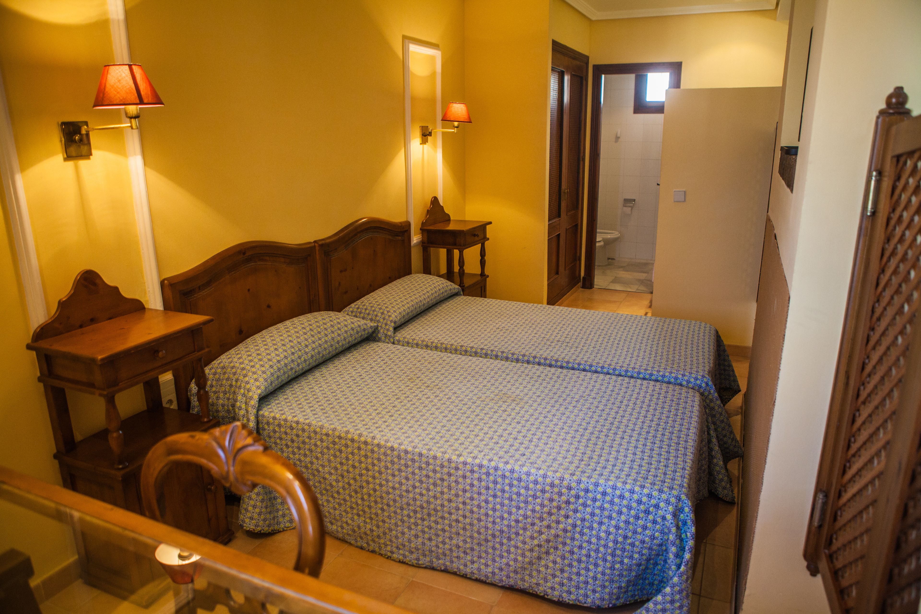 Una habitación de hotel con dos camas, cabecero de madera, mesita de noche, lámpara y espejo.