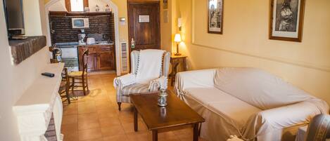 A living room with a sofa, armchair, and a small table. There is a kitchen area with a microwave and a door.