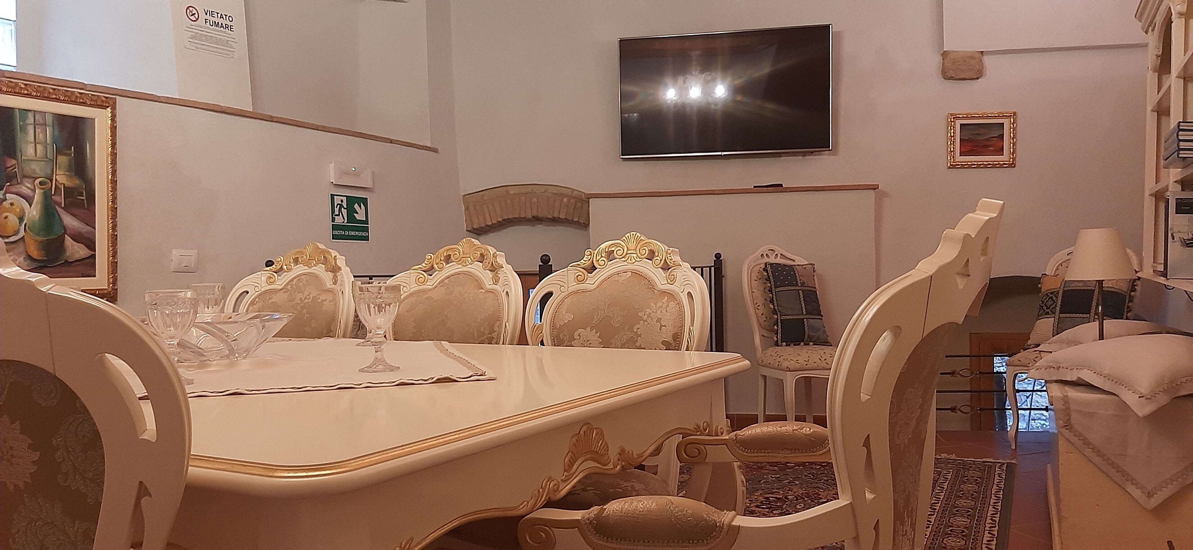 Un área de comedor con mobiliario blanco ornamentado, un televisor de pantalla plana en la pared y una obra enmarcada sobre la mesa.