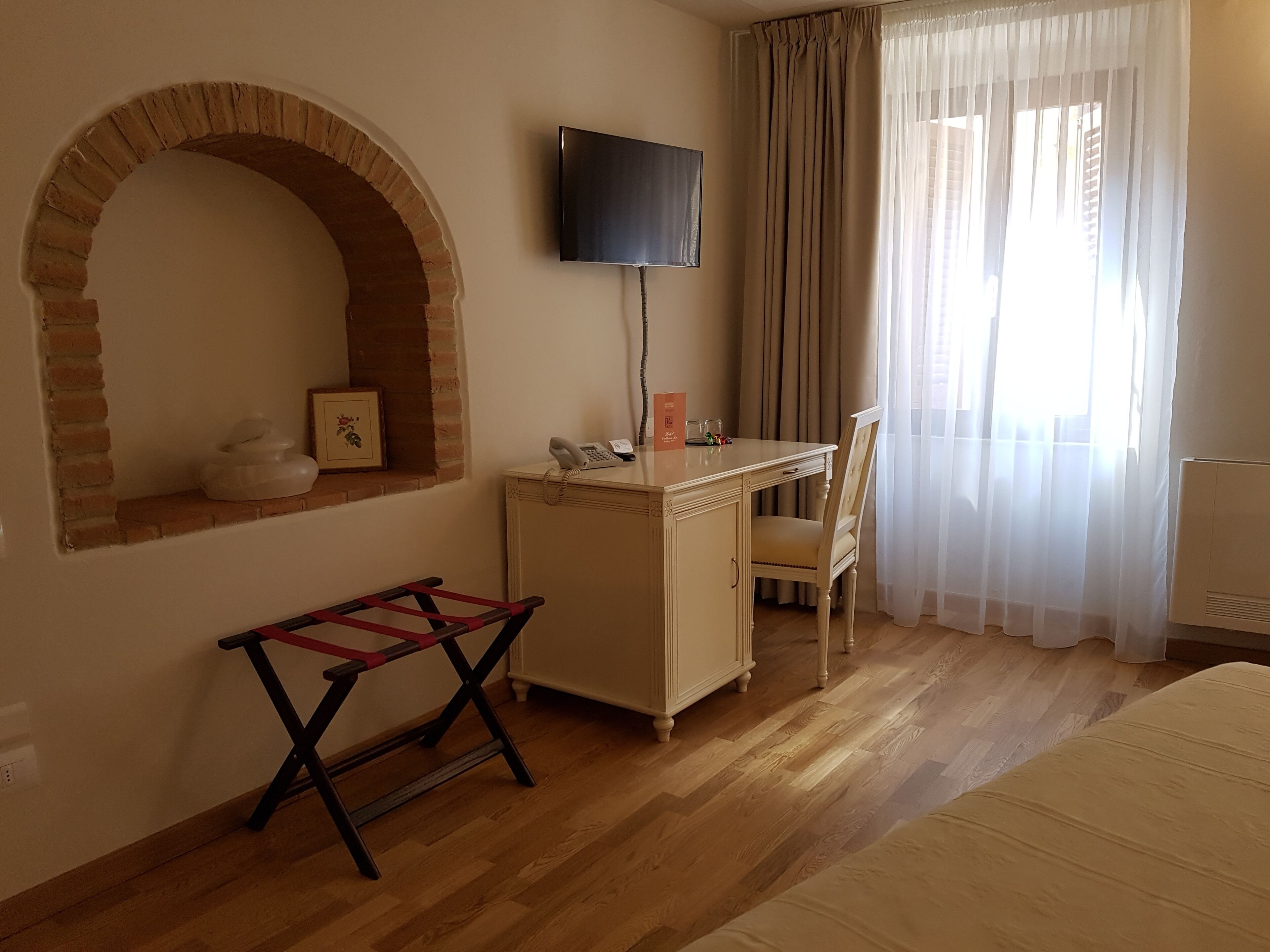 A room with a brick archway, a desk with a telephone, a chair, a TV mounted on the wall, and a window with sheer curtains.