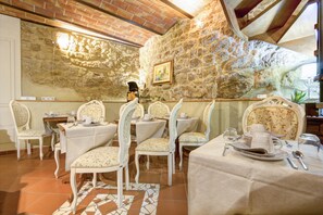Un comedor con paredes de piedra, sillas blancas tapizadas y mesas dispuestas para una comida.