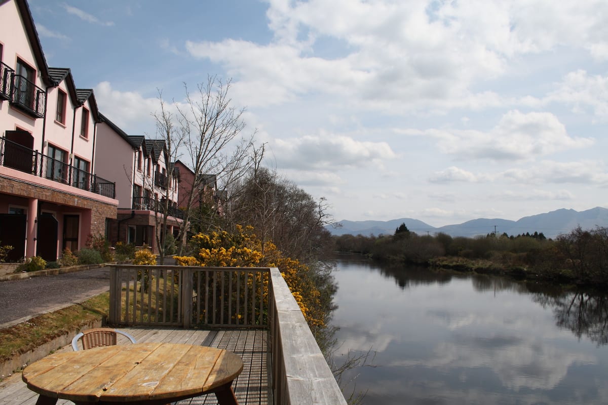 A riverside view with a wooden table and chairs, a wooden fence, and buildings with balconies.