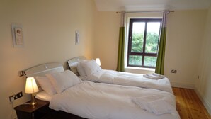 Una cama doble con ropa de cama blanca, una mesita de noche con lámpara, una ventana con cortinas y suelo de madera.