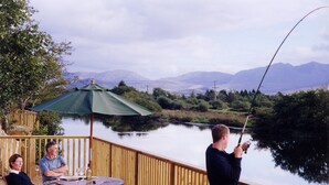 Una terraza de madera con una mesa y sillas, con vista a un lago tranquilo y montañas al fondo.