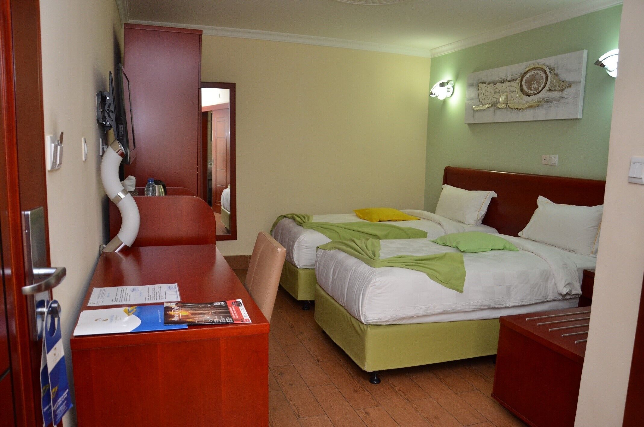 A hotel room with a desk, two beds, a mirror, and a wall-mounted artwork.