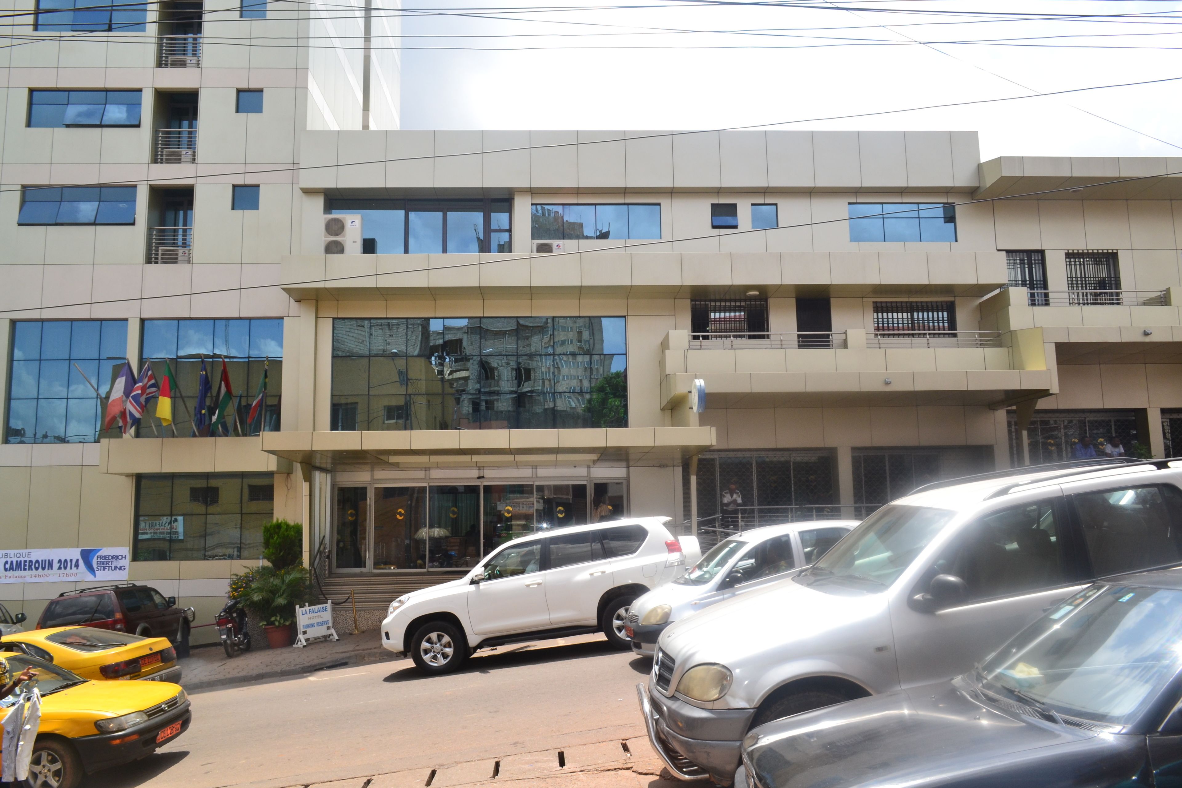 Un edificio de varios pisos con un diseño moderno, que cuenta con ventanas y balcones amplios. Varios coches están estacionados en la parte delantera, y hay banderas exhibidas en el exterior.