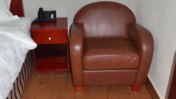 Un dormitorio con cama, una butaca de cuero marrón, una mesita de noche de madera con teléfono y suelo de madera.