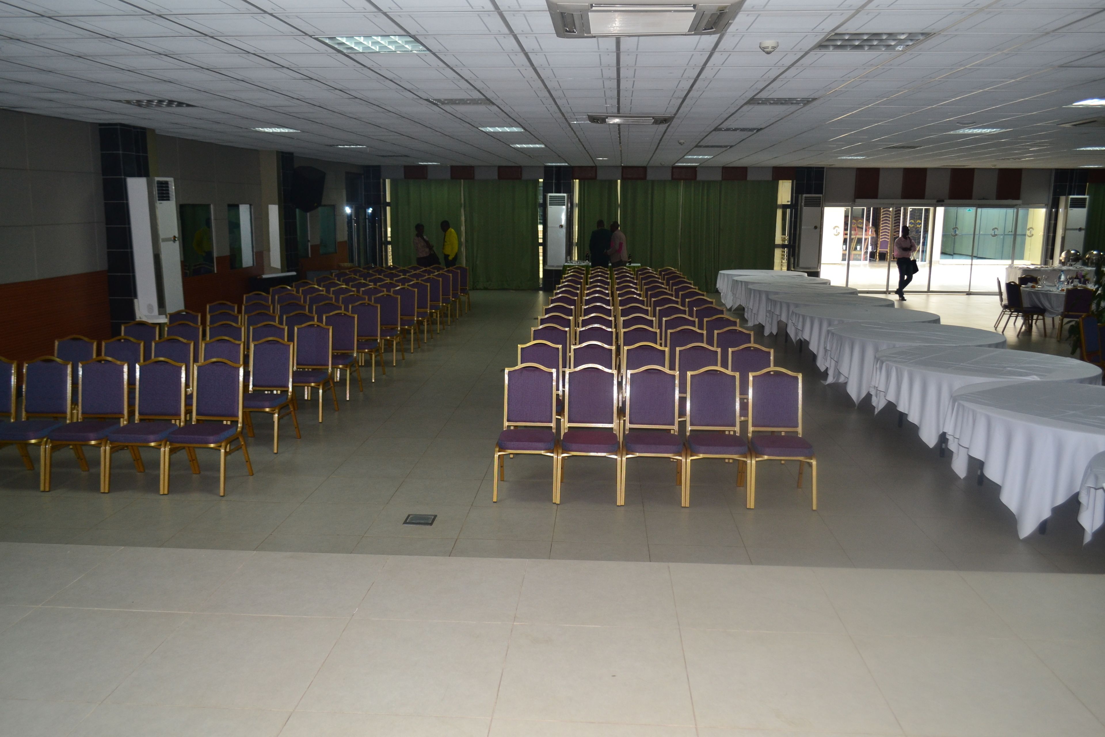 A conference room with rows of purple chairs and white tables, large windows, and a green curtain backdrop.