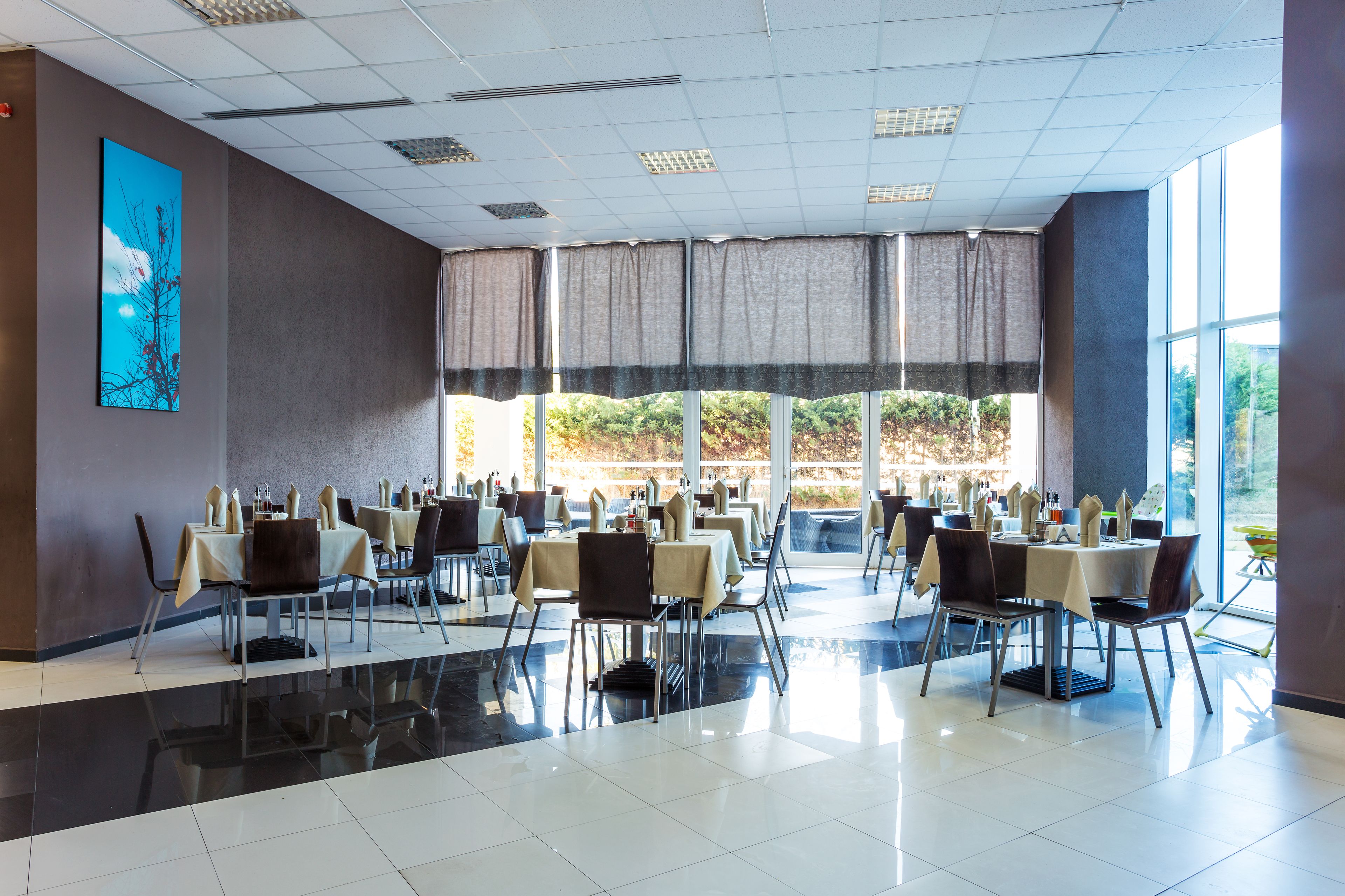 Un comedor amplio con varias mesas y sillas, ventanas grandes con cortinas y suelo de baldosas.