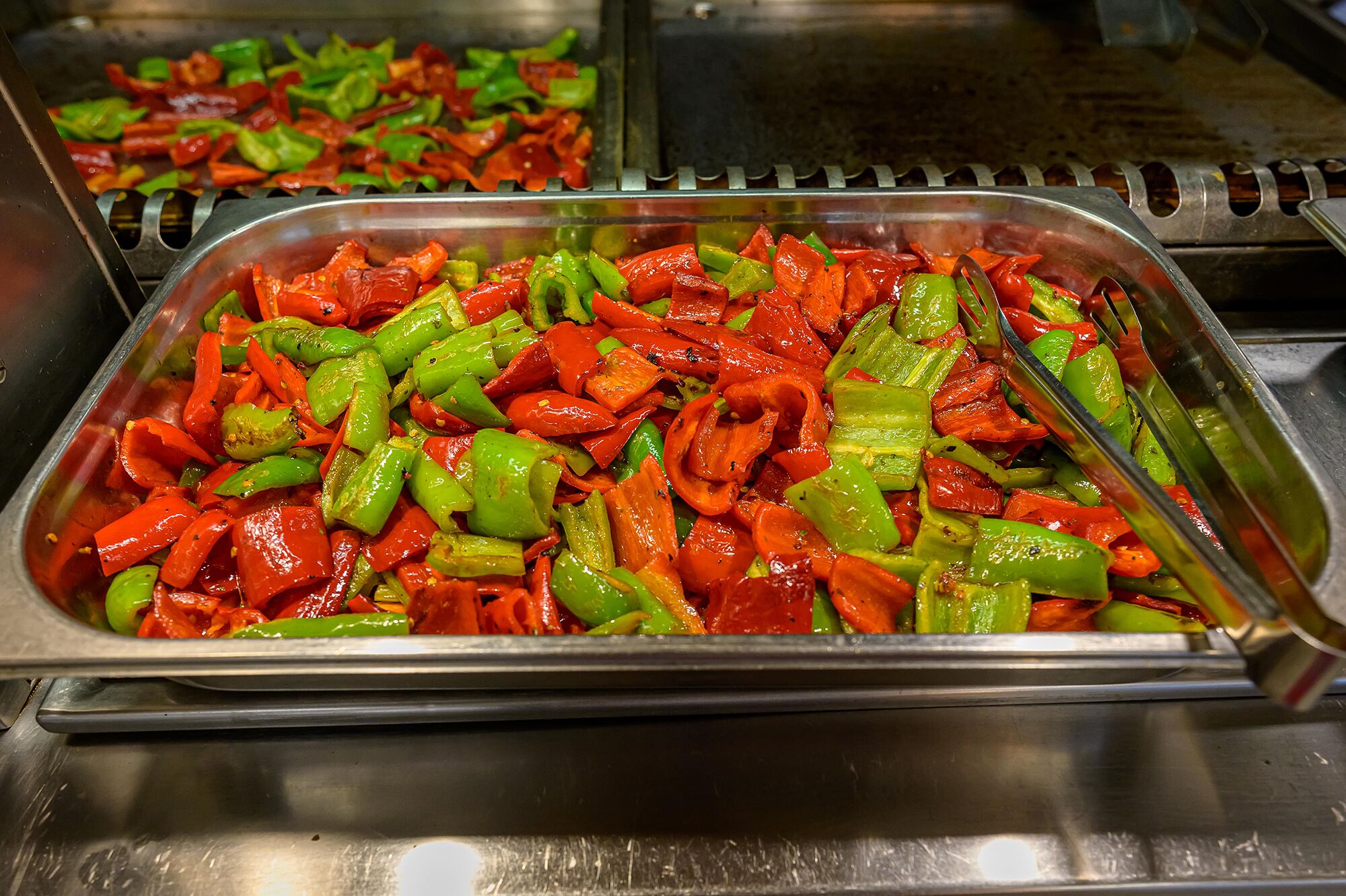 A buffet with a variety of sliced vegetables, including red and green bell peppers, arranged in metal trays.