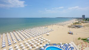 A beach with rows of sun loungers and umbrellas, a swimming pool, and a distant view of the sea and buildings. - Blue Pearl Hotel (Sunny Beach)