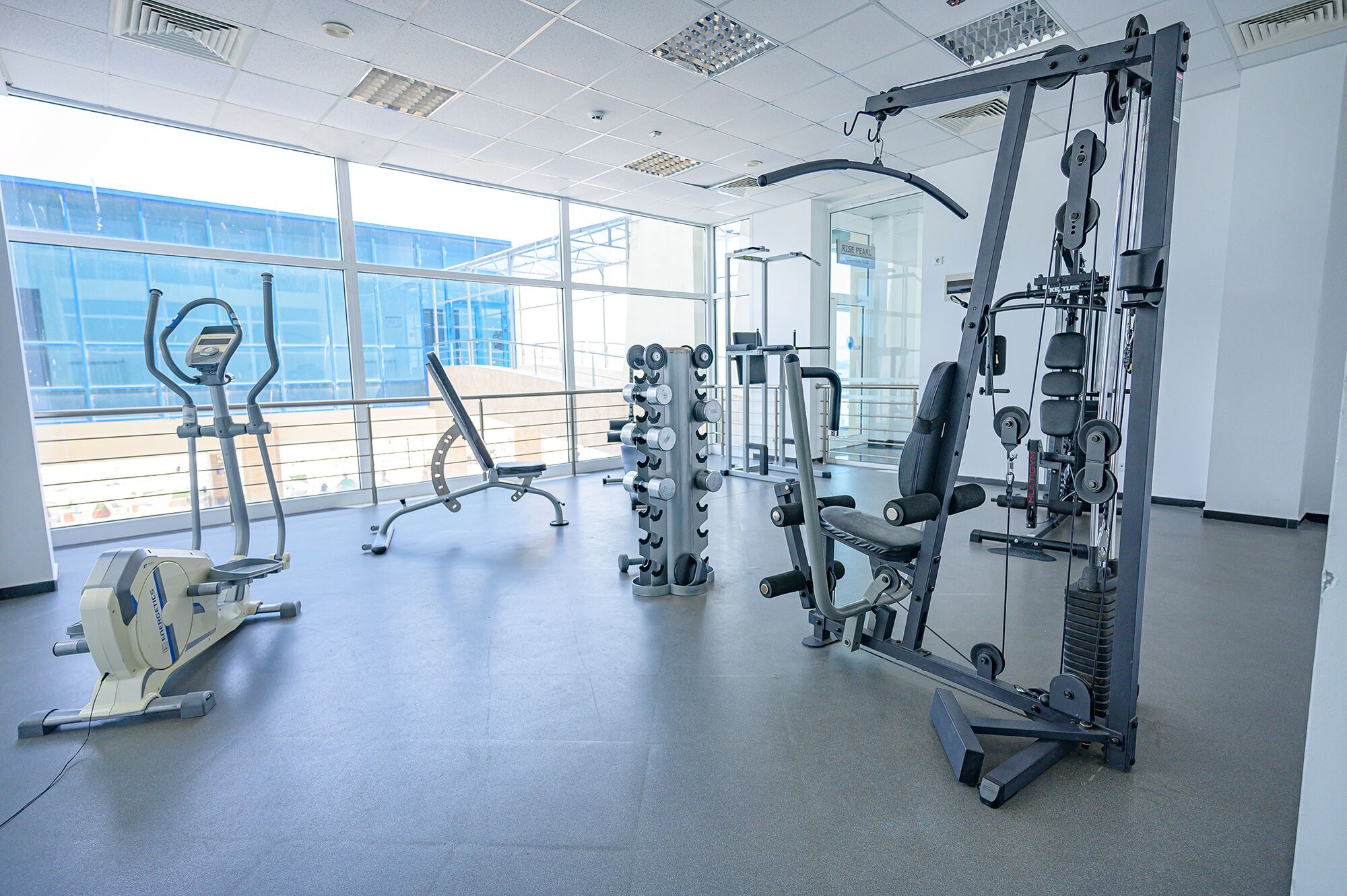 A gym with various exercise equipment, large windows, and a view of a building outside.