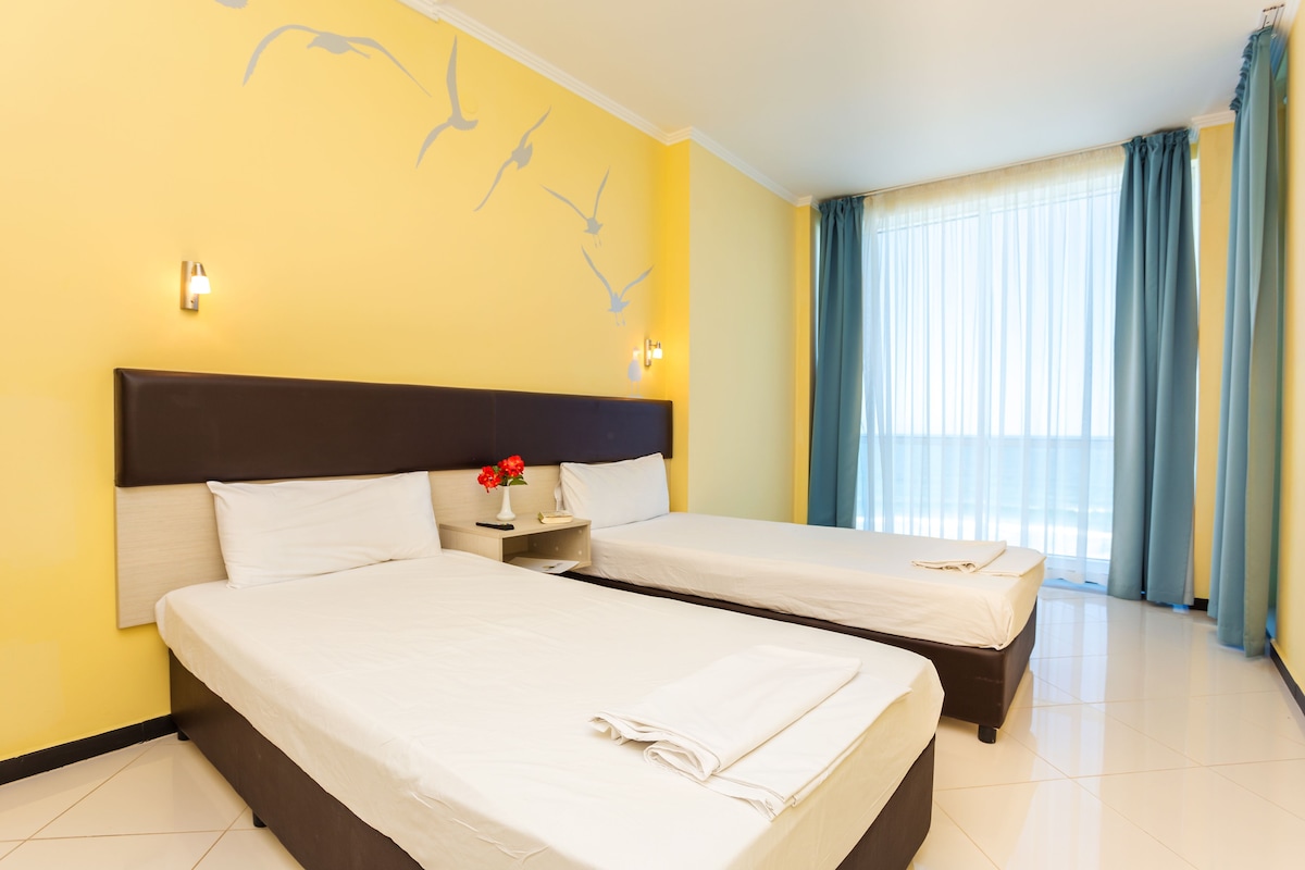 A hotel room with two beds, a nightstand with a vase of flowers, and a window with curtains.