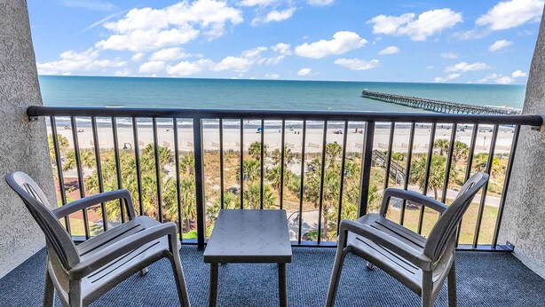 Un balcón con dos sillas y una mesa, con vistas a una playa y un muelle.