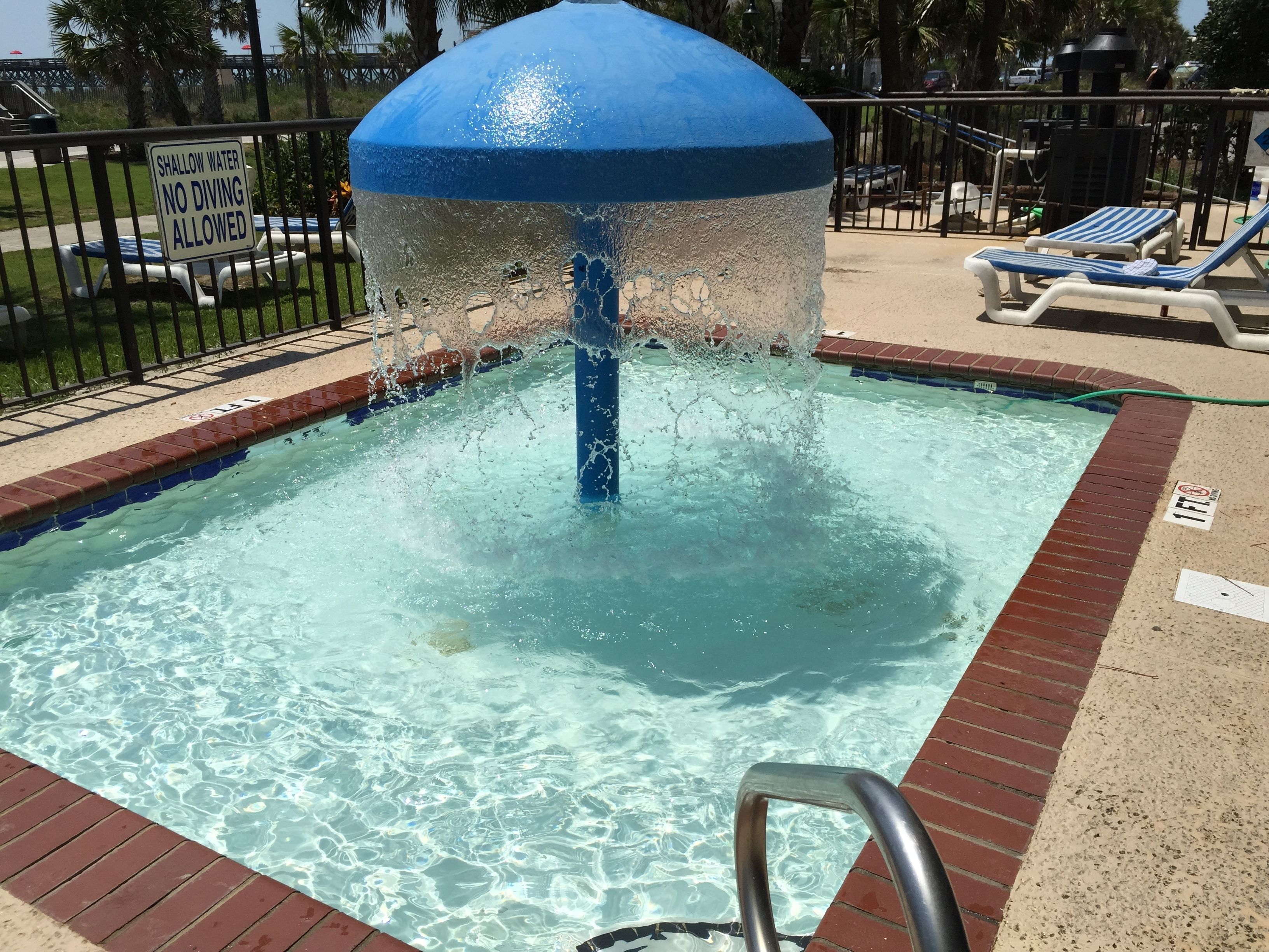 Une piscine avec une fontaine d’eau bleue, un panneau indiquant la faible profondeur de l’eau et un panneau interdisant la plongée.