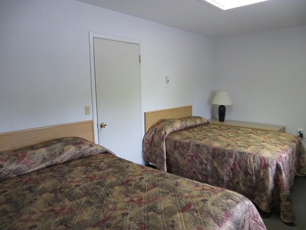 A room with two beds, a door, a nightstand with a lamp, and a ceiling light.