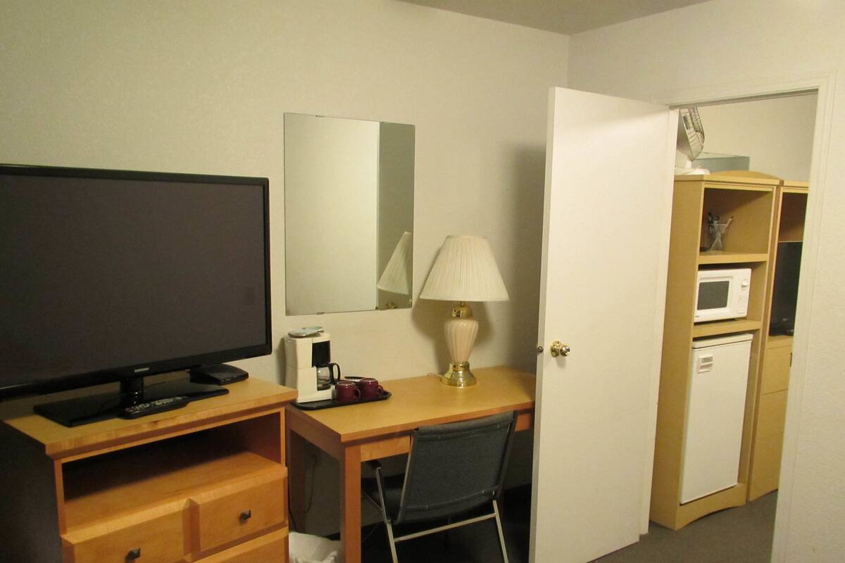 Habitación de hotel con televisor, escritorio, silla, lámpara, cafetera, microondas y refrigerador pequeño.
