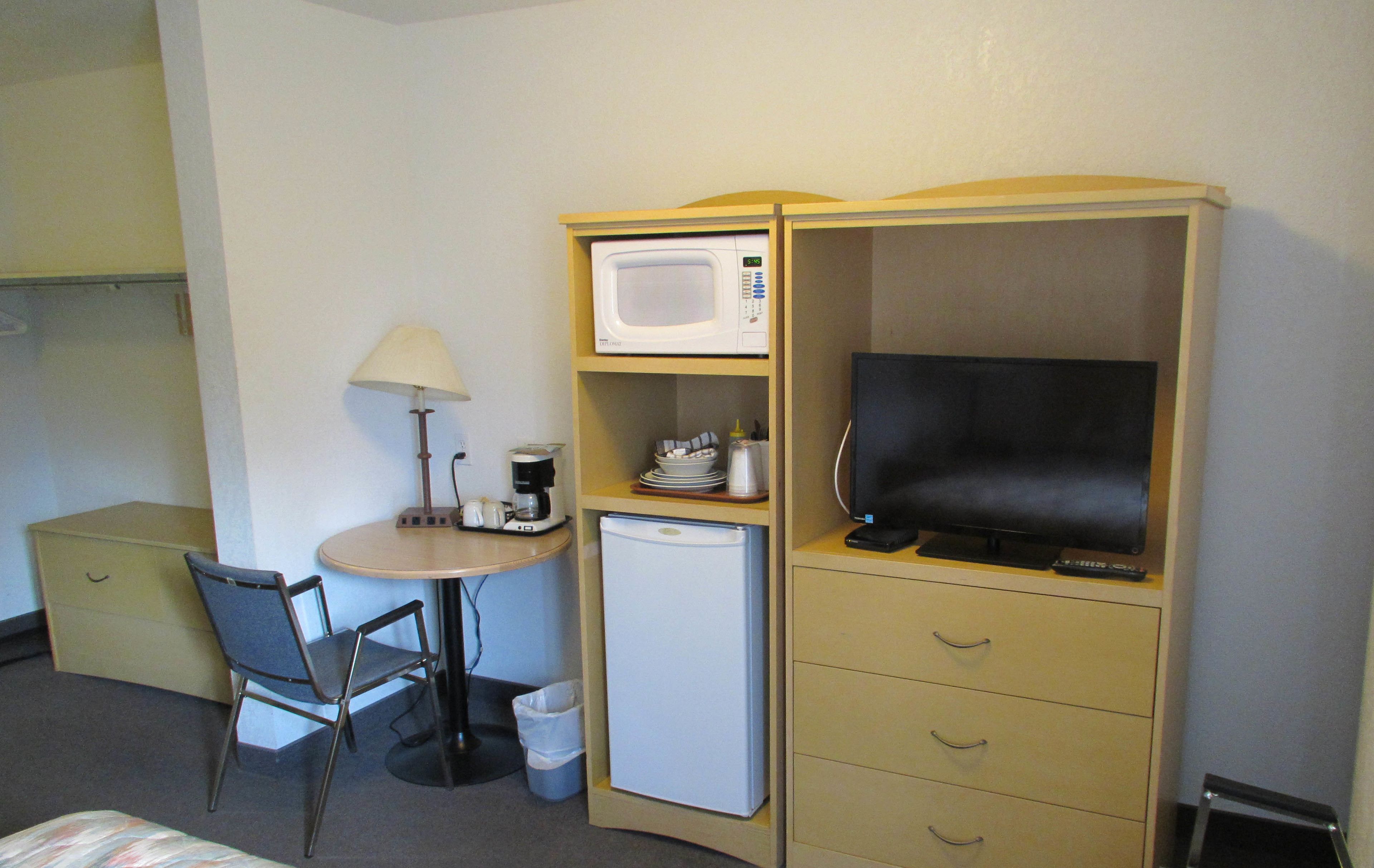 A compact hotel room with a bed, a desk with a lamp, a microwave, a small refrigerator, a television, and a chair.