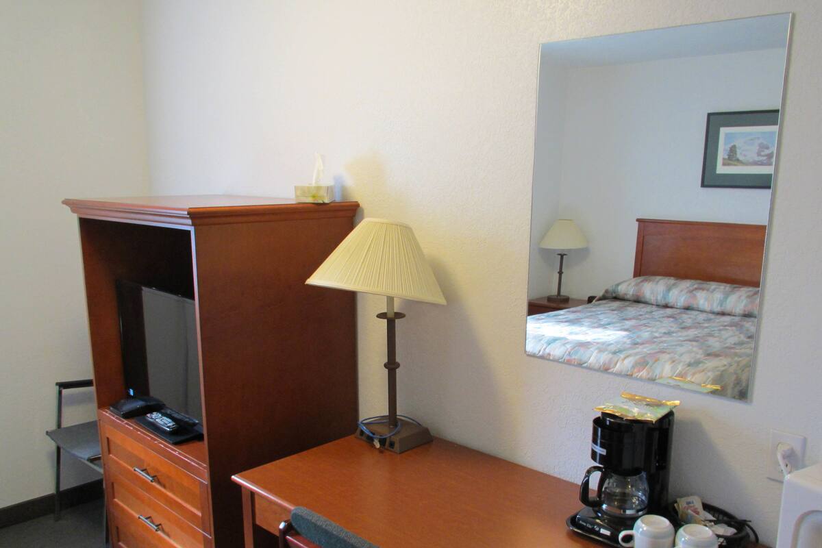 Una habitación de hotel con un aparador de madera, una lámpara, una cama con colcha estampada, una cafetera y un espejo que refleja la cama.