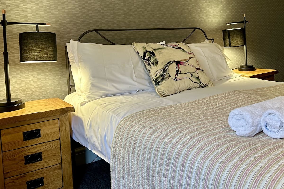 A neatly made bed with white linens, a patterned throw, and rolled towels on the side. There are two bedside tables with lamps and a wooden nightstand with drawers.