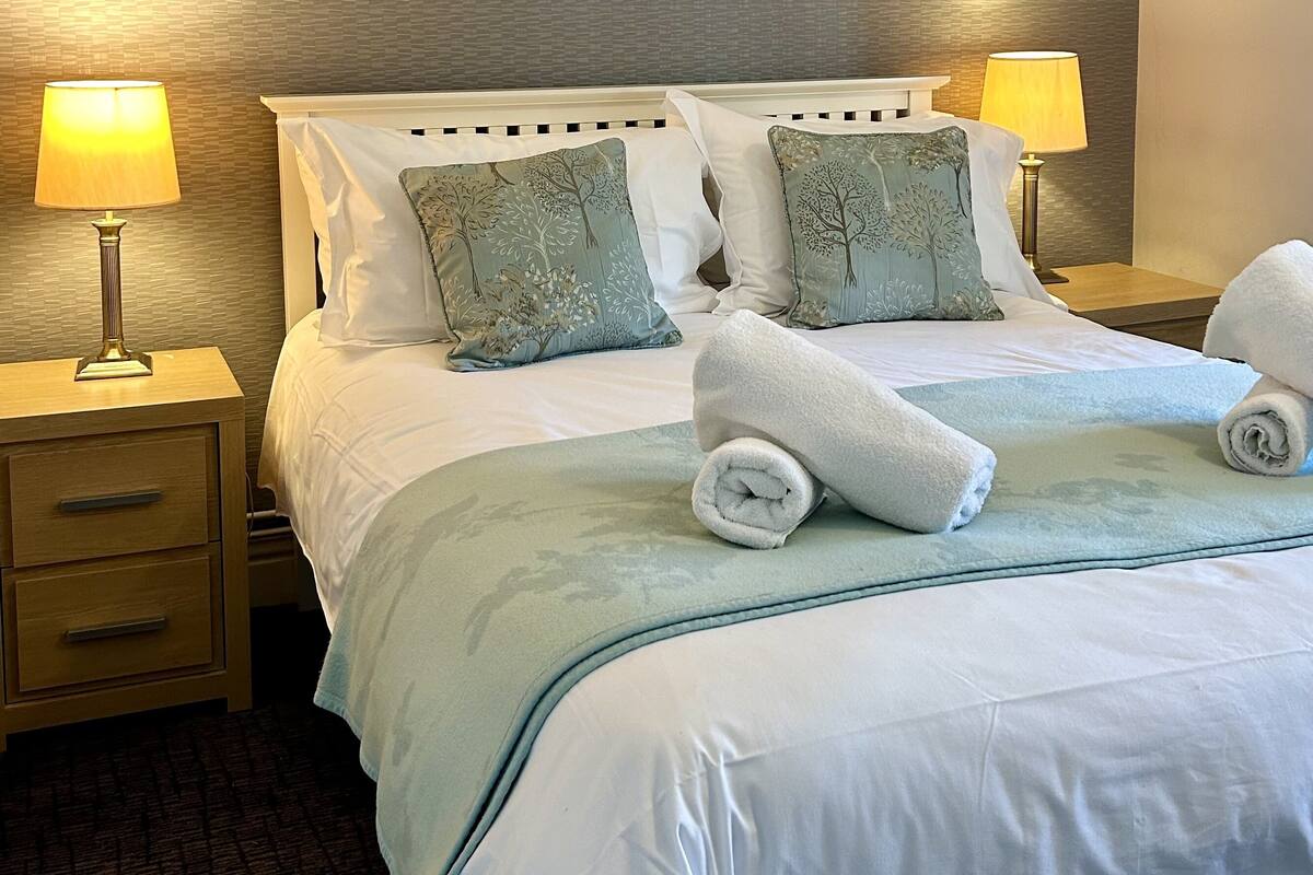 A neatly made bed with white linens, two pillows with a tree pattern, and two rolled towels. There are two bedside tables with lamps, one on each side of the bed.