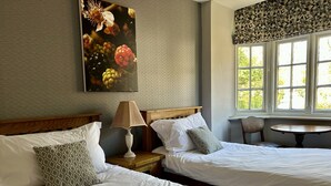 A hotel room with two beds, a nightstand with a lamp, a chair, and a window with a view of greenery. - The Talbot (Blandford Forum)
