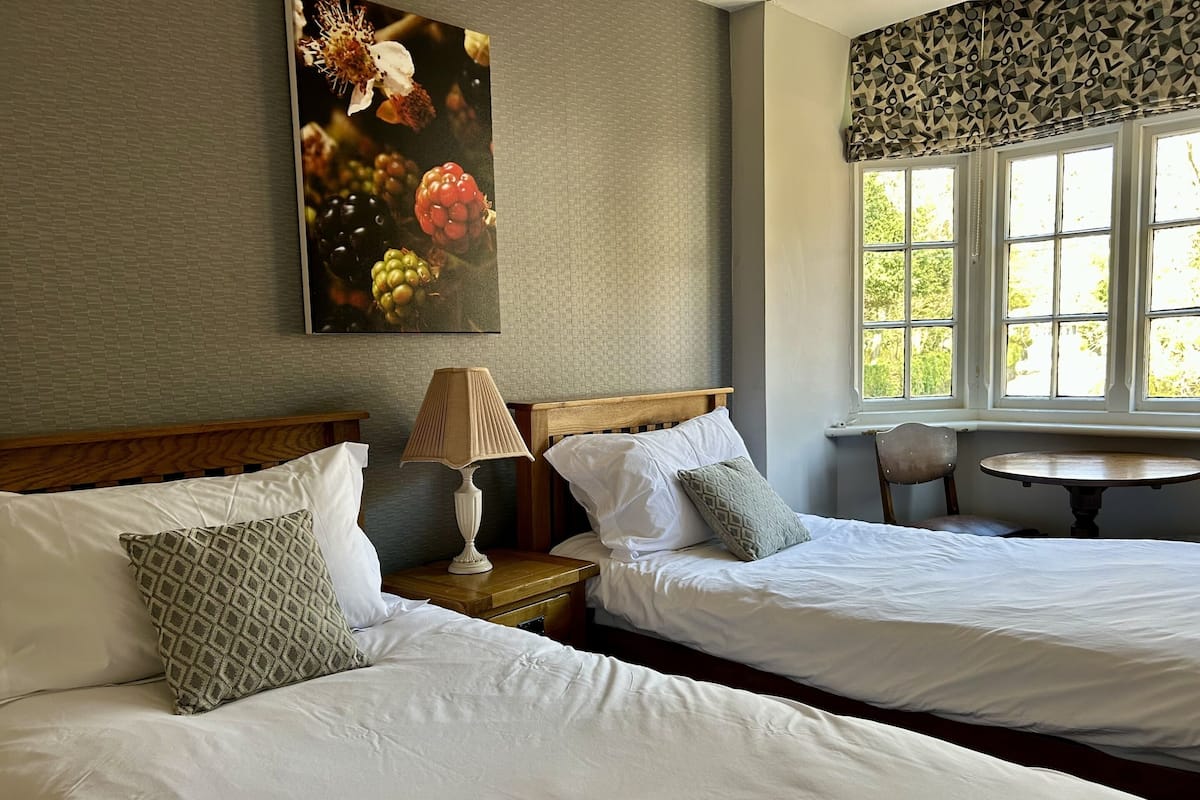 A hotel room with two beds, a nightstand with a lamp, a chair, and a window with a view of greenery.