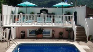 A swimming pool with a white deck, a small set of stairs leading to a covered area with umbrellas, and a view of hills in the background. - Windsor Motel (Lake George)