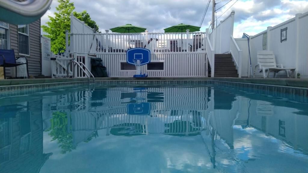 A swimming pool with a basketball hoop, white fence, and a covered area with chairs.