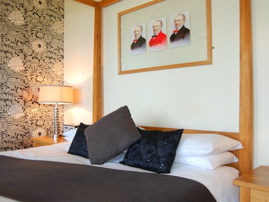 A neatly made bed with a dark blanket and pillows, a wooden headboard, a wall with floral wallpaper, and a framed portrait above the bed.
