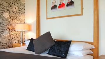 A neatly made bed with a dark blanket and pillows, a wooden headboard, a wall with floral wallpaper, and a framed portrait above the bed.