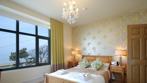 A bedroom with a large window, a wooden bed, two bedside tables with lamps, and a door. - The Gladstone (Penmaenmawr)