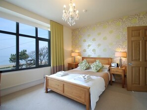 A bedroom with a large window, a wooden bed, two bedside tables with lamps, and a door. - The Gladstone (Penmaenmawr)