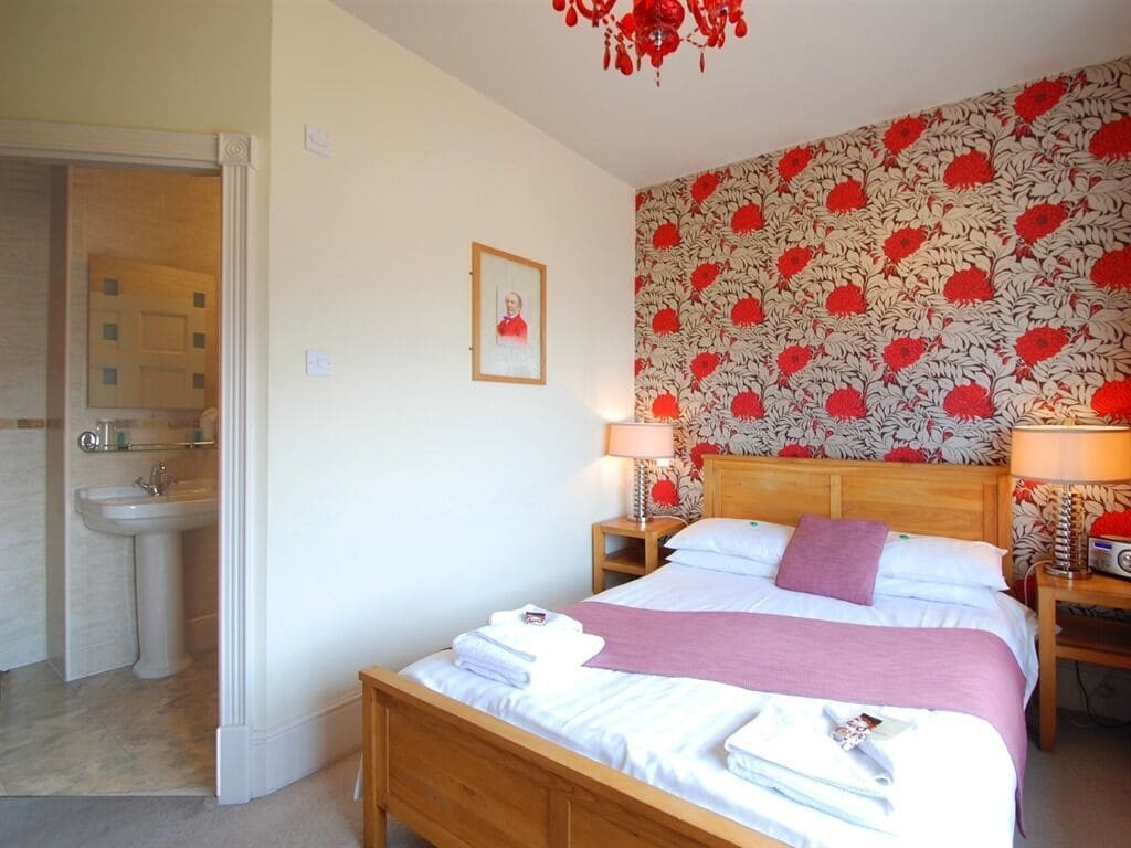 A bedroom with a bed, bedside tables, a nightstand, a lamp, a framed picture on the wall, and a bathroom with a sink and mirror.