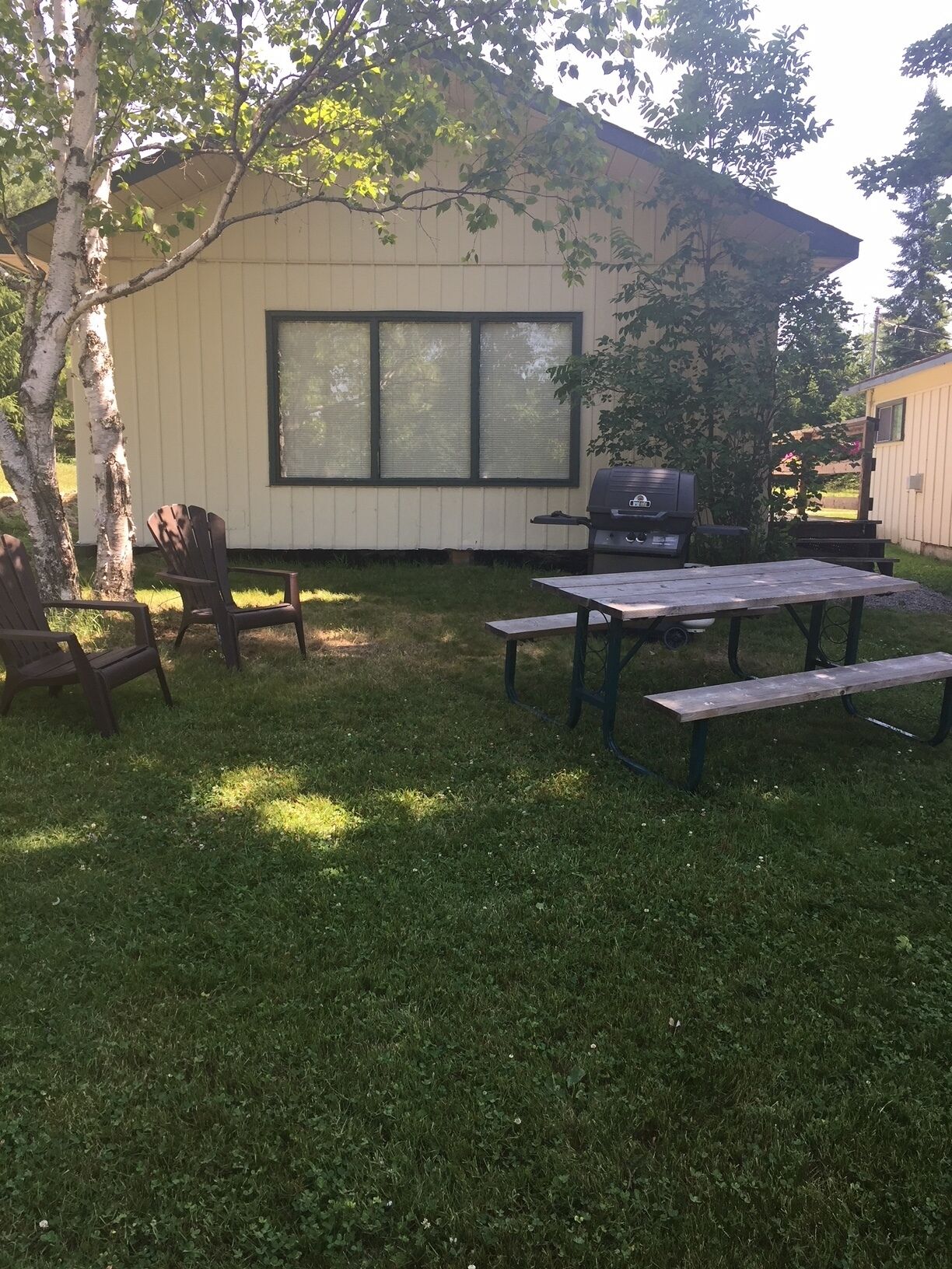 A backyard with a wooden table and chairs, a grill, and a house with large windows.