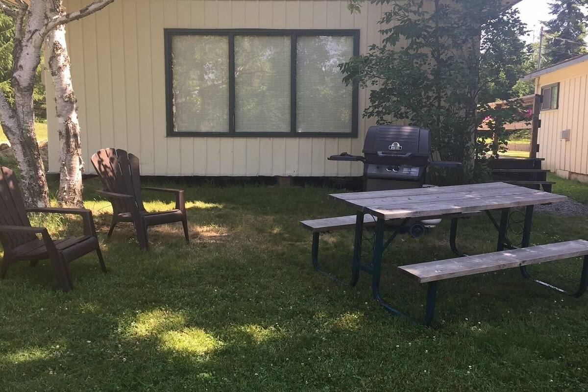 A backyard with a wooden table and chairs, a grill, and a house with large windows.
