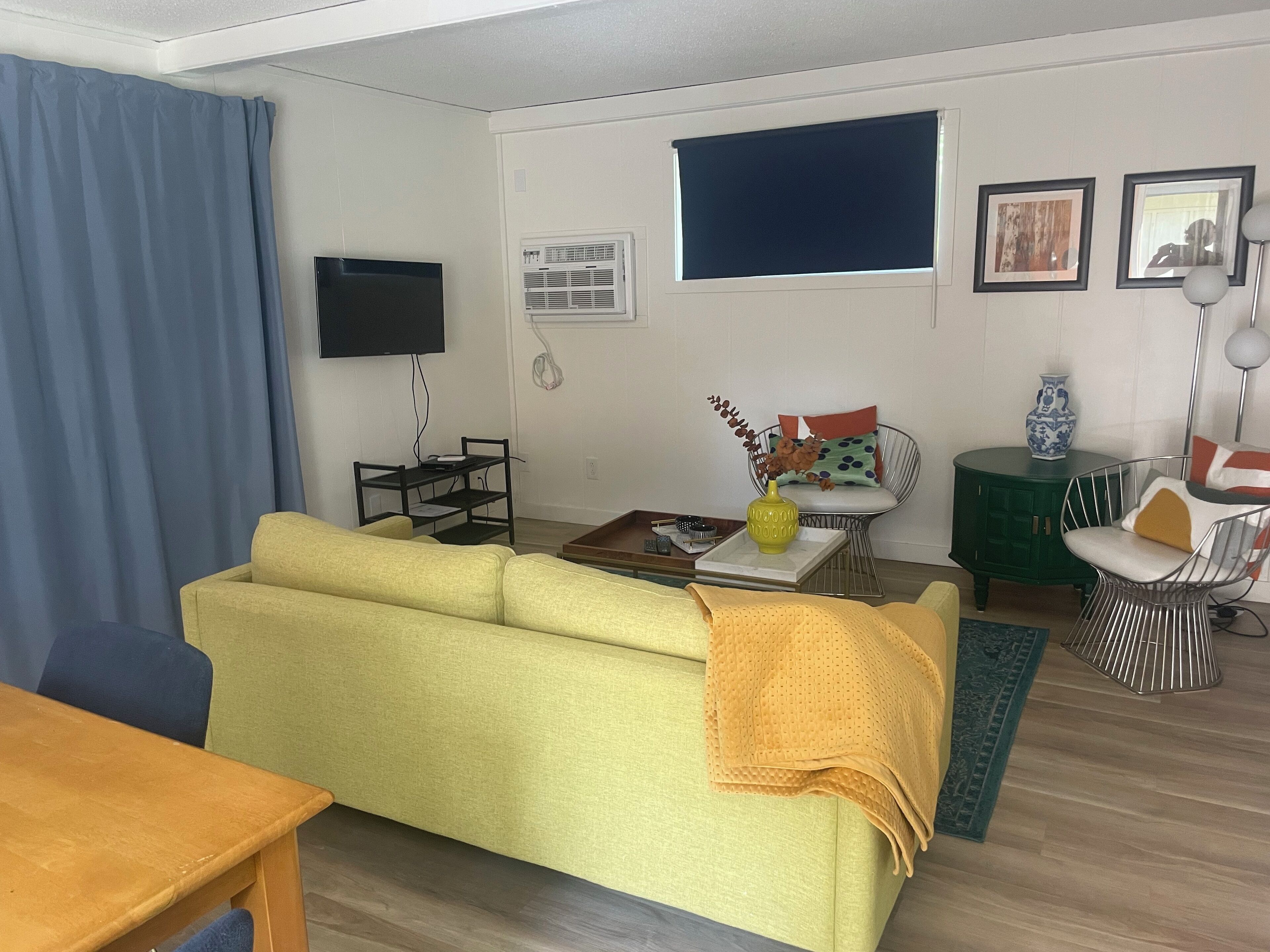 Ein Wohnzimmer mit gelber Couch, Couchtisch, Fernseher, kleinen Beistelltisch und einem grünen Schrank.