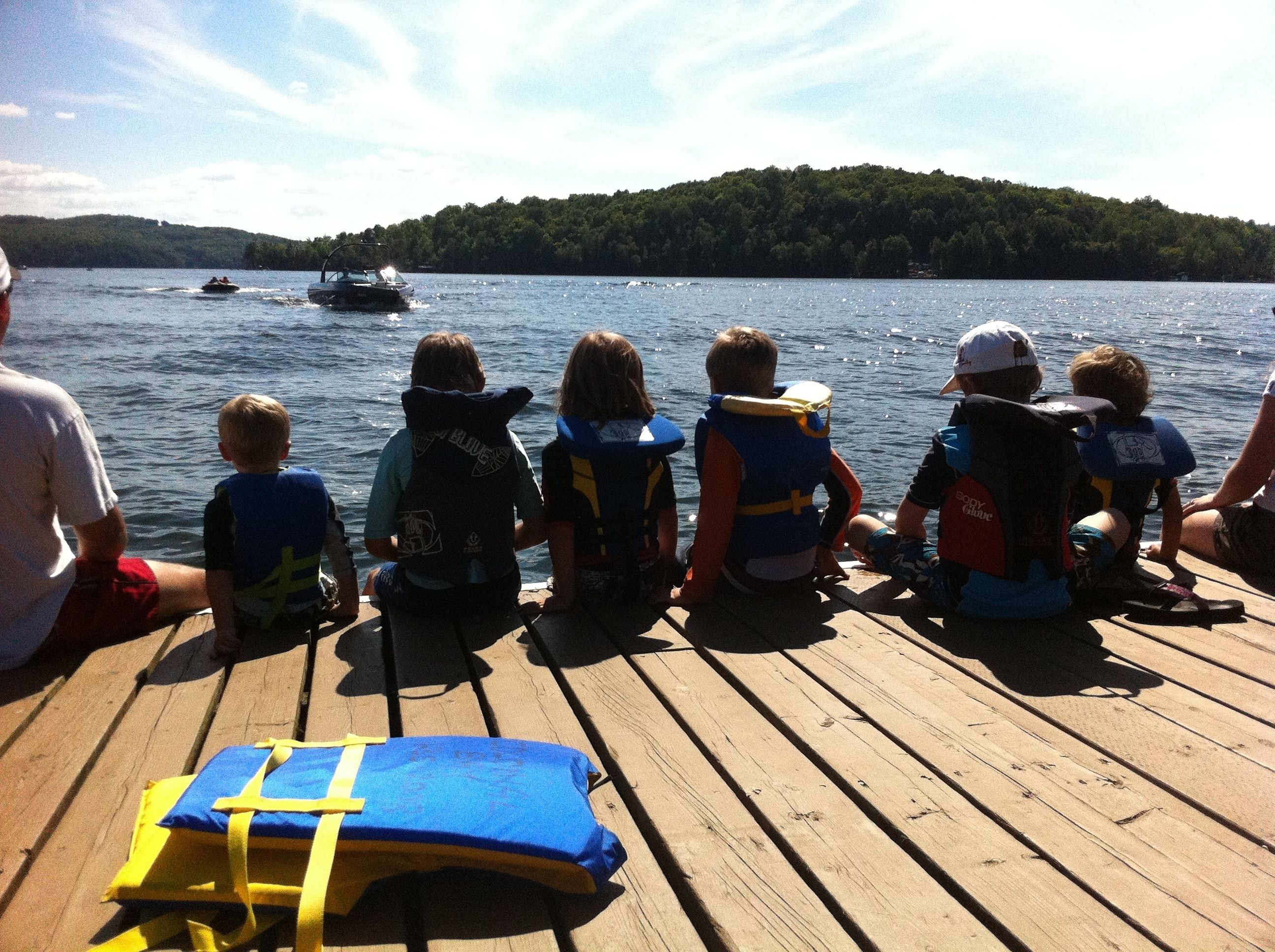 Eine Gruppe Menschen auf einem hölzernen Steg mit Blick auf einen See, in der Ferne Boote und ein bewaldeter Hügel.
