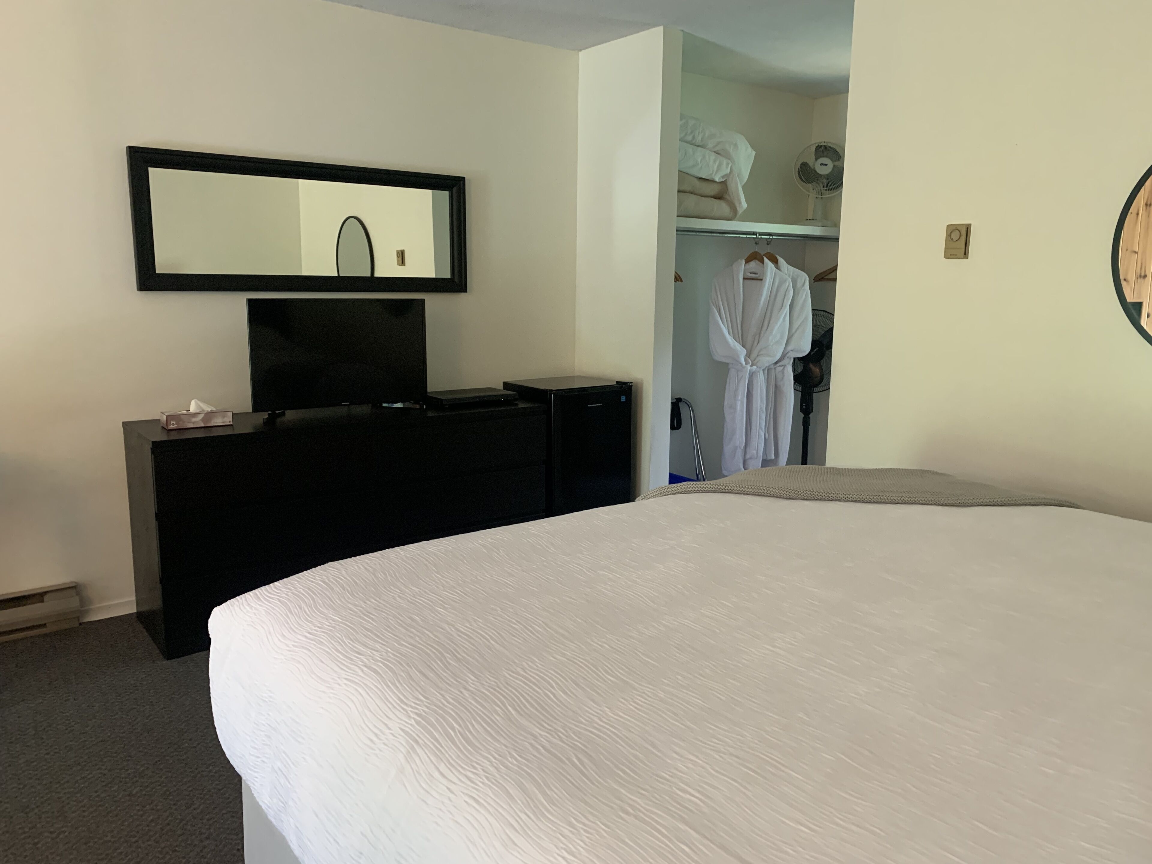 Et hotelværelse med en stor seng, et fjernsyn på en kommode, et spejl, et skab med badekåbe og en ventilator.