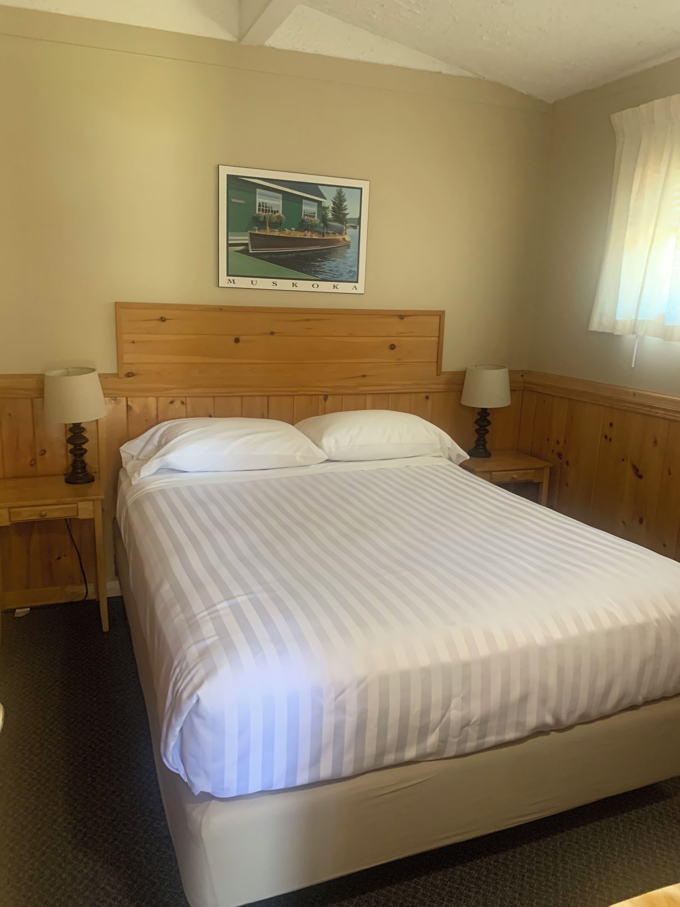 Een netjes opgemaakt bed met een gestreept dekbedovertrek, houten hoofdbord en twee bedlampjes. Een lijst met een foto van een huis hangt boven het bed, met daarop de tekst 
