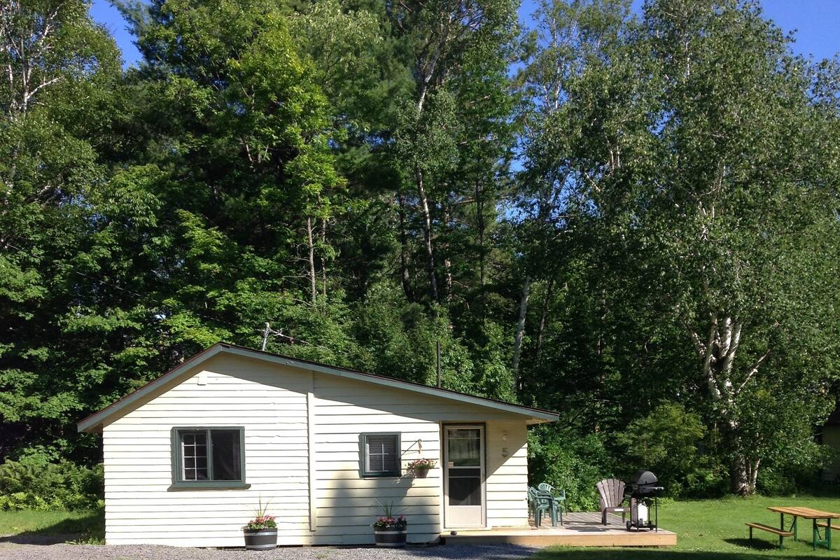 A small, single-story cabin with a porch, surrounded by a grassy area and trees.