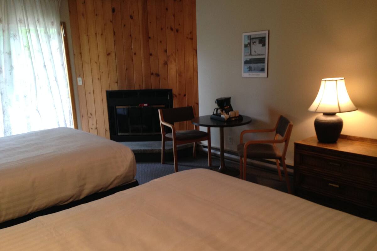 A hotel room with two beds, a small table with a lamp, a chair, and a fireplace.