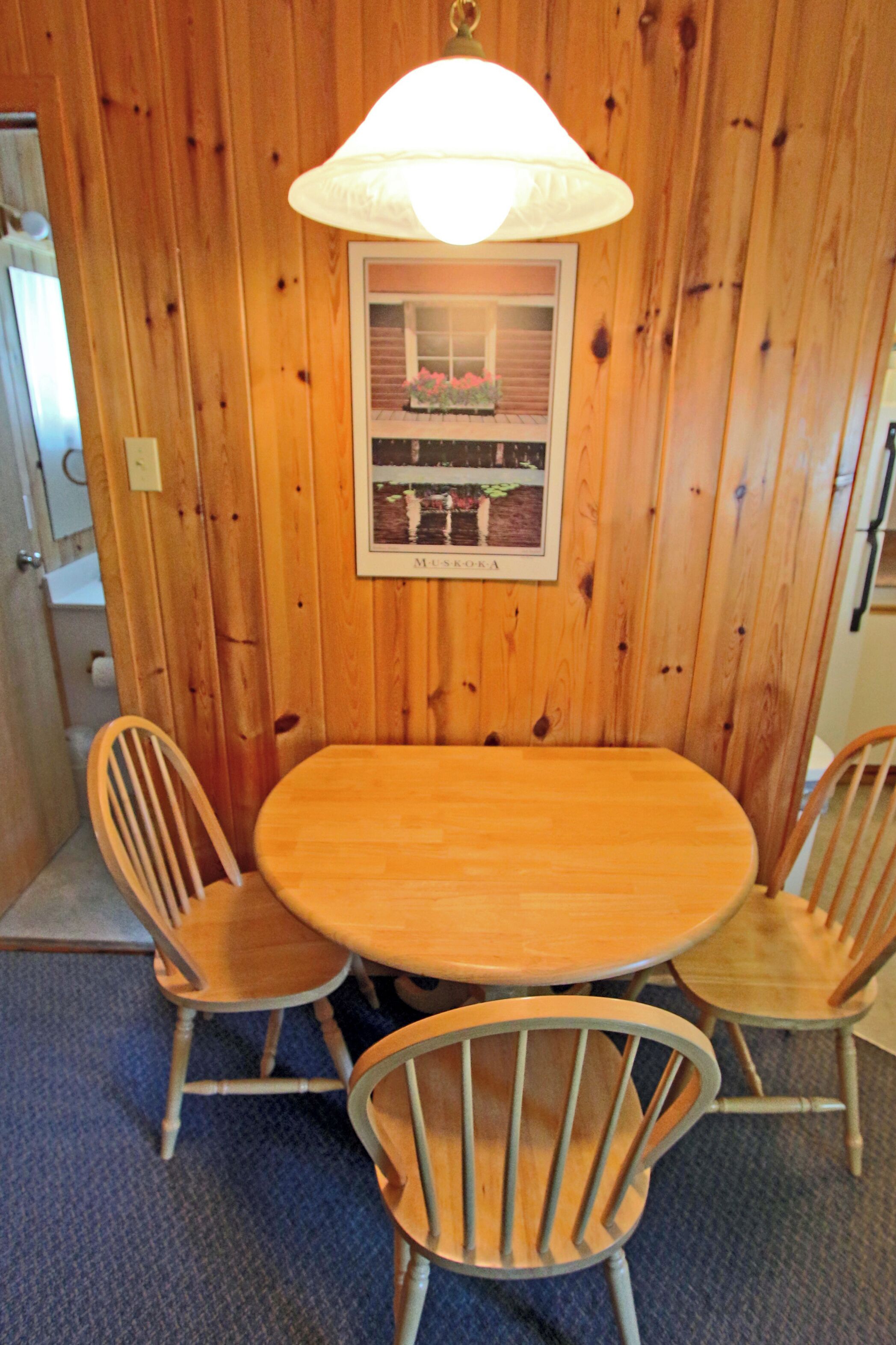 Une pièce aux murs lambrissés, avec une table ronde et quatre chaises. Un tableau encadré est accroché au mur, au-dessus de la table.
