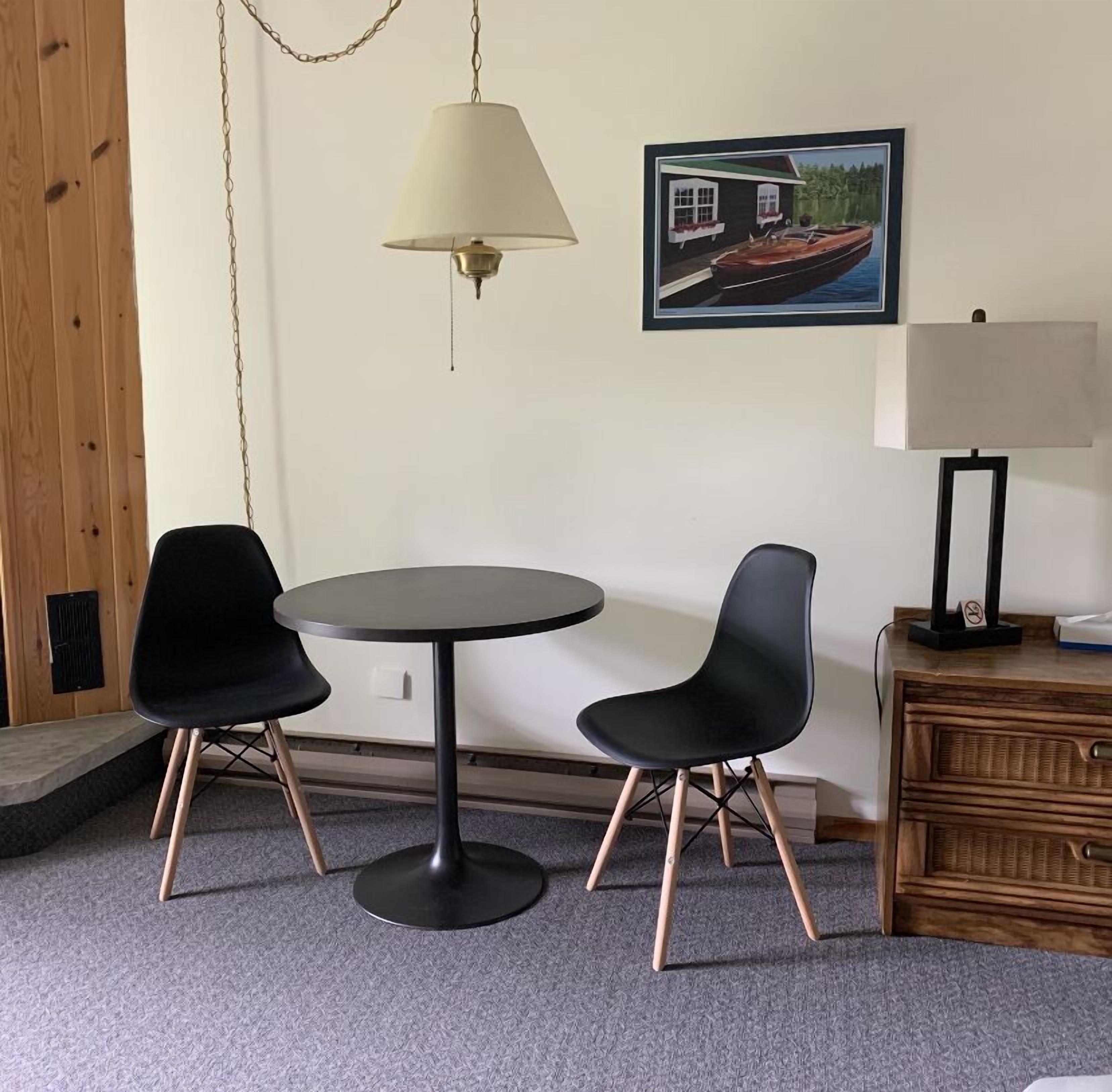 Una habitación con una mesa redonda, dos sillas negras, una lámpara, un cuadro con una imagen de un barco y un armario de madera.