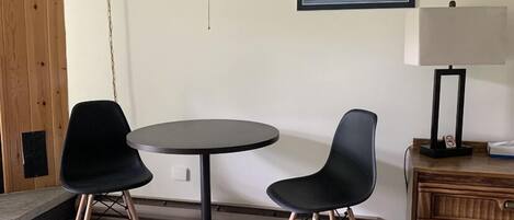 Una habitación con una mesa redonda, dos sillas negras, una lámpara, un cuadro con una imagen de un barco y un armario de madera.