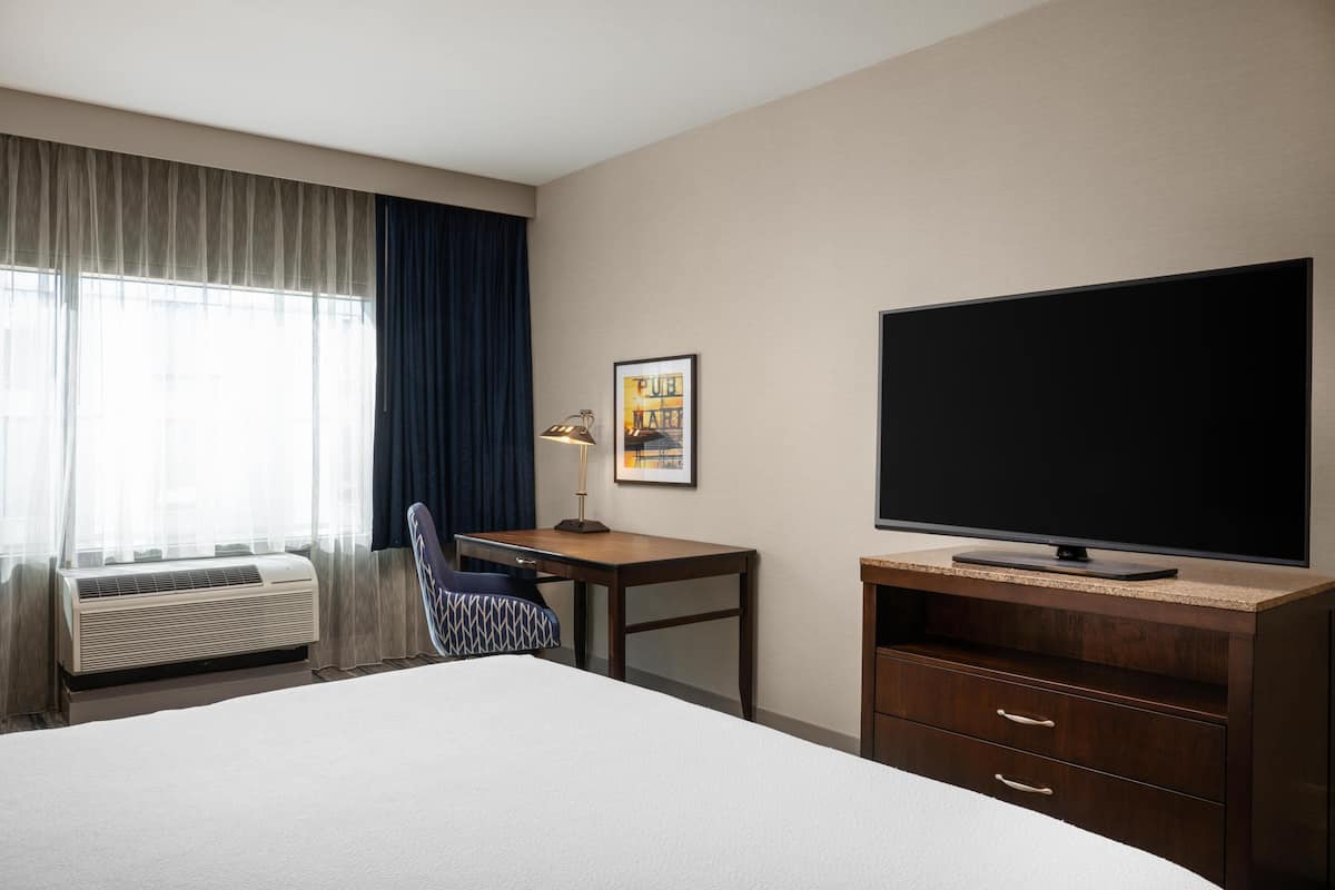 A hotel room with a bed, desk, chair, television, and air conditioning unit.